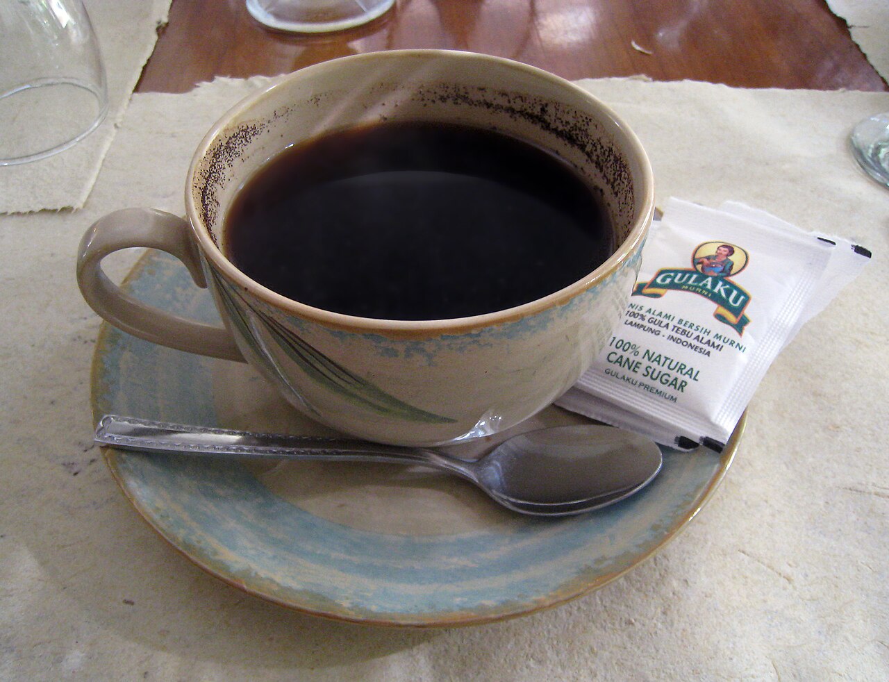 Javanese tubruk with grounds visible around the inner cup. The sugar packet is a serving choice, not proof of sugar already added.