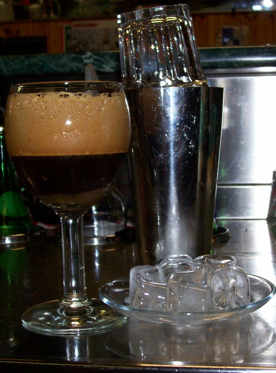 A source-identified caffè shakerato beside a shaker and ice. The photograph is a finished-drink presentation; exact coffee and sugar amounts are not visible.